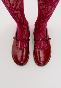 Zapatos Mary Jane de cuero rojo con punta redonda y tacón plano, que cuentan con una correa con hebilla. Combinados con medias fucsias con un patrón de encaje.