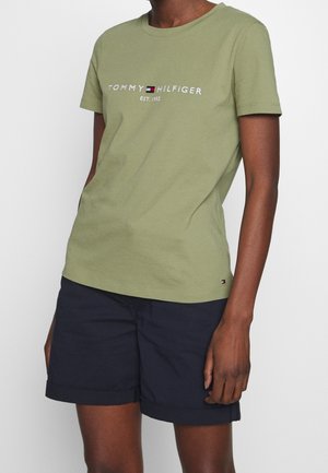 Persoon draagt een olijfgroen Tommy Hilfiger T-shirt met logo en marineblauwe shorts met opgerolde zoom tegen een effen achtergrond.