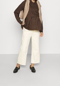 Chemise à manches longues à carreaux marron et noir, gilet beige sans manches, pantalons larges blancs, et mocassins noirs avec des détails dorés.