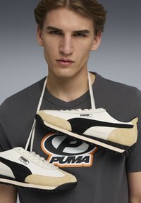 PUMA Easy Rider schoenen met witte leren en zwarte suède accenten; ronde neus, zwarte rubberen zool en veters; gedragen om de nek.