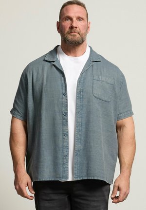 Bärtiger Mann, der ein lockeres, kurzärmeliges graues Hemd über einem weißen T-Shirt trägt und vor einem schlichten Hintergrund steht.