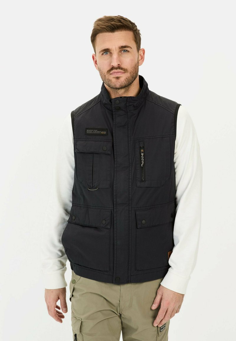 Schwarwest aus strapazierfähigem Material, mit mehreren Taschen, Reißverschlussdetails und Stehkragen. Getragen über einem weißen Langarmhemd.