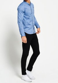 Jasnoniebieska koszula zapinana na guziki z kieszonką z przodu, w połączeniu z czarnymi spodniami slim fit i białymi trampkami. Koszula ma teksturowany materiał.