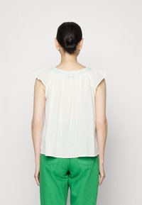 Blusa branca sem mangas com ombros franzidos, decote redondo bordado e tecido leve e arejado. Combinada com calças verdes.