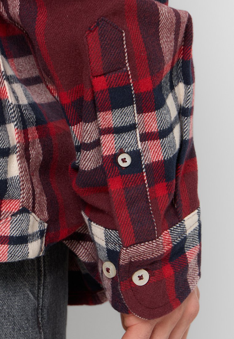 Camicia in flanella a quadri rossa e blu navy con polsini bottonati. Nota per i contrasti di texture e colori, con accenti e cuciture bianchi.