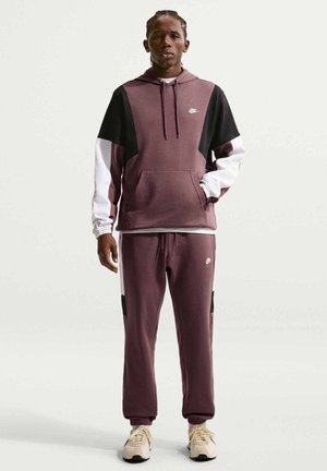 Jeune homme portant un sweat à capuche Nike bordeaux, noir et blanc avec un pantalon de jogging assorti et des baskets beige, debout devant un fond uni.