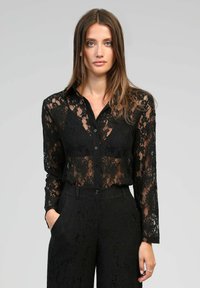 Blouse en dentelle noire avec manches longues, fermeture à boutons et motif floral complexe, associée à un pantalon en dentelle noire assorti à la coupe ajustée.