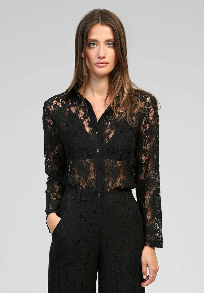 Blouse en dentelle noire avec manches longues, fermeture à boutons et motif floral complexe, associée à un pantalon en dentelle noire assorti à la coupe ajustée.