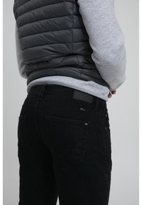 Jeans de mezclilla negros con corte slim, que cuentan con bolsillos traseros y una superficie texturizada, combinados con una sudadera gris y un chaleco acolchado.