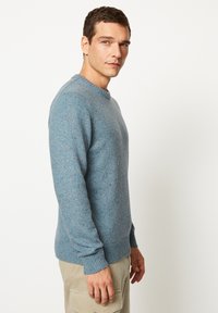 Blau gestrickter Pullover mit Rundhalsausschnitt, der eine gesprenkelte Textur und gerippte Bündchen sowie Saum aufweist. Das Model trägt hellfarbene Hosen.