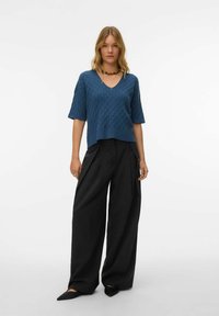 Tirkizno plavi džemper s V-izrezom i teksturnim uzorkom, uparen s visokim crnim širokim hlačama i šiljastim crnim cipelama.