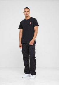 Tricou negru din bumbac cu o grafică mică de cartofi prăjiți roșii și galbeni, asociat cu pantaloni cargo negri cu buzunare cu fermoar și adidași albi.