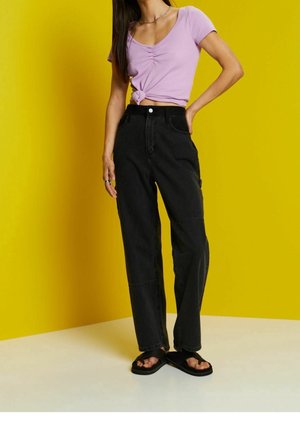 Vrouw draagt een lila, geknoopte top met korte mouwen, een zwarte broek met hoge taille en zwarte sandalen, staand voor een gele achtergrond.