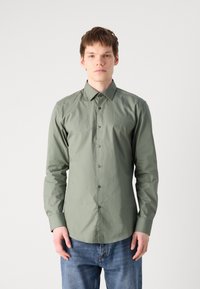 Cămașă lungă, verde deschis, cu guler îmbrăcat cu nasturi, având o textură netedă și nasturi închis la culoare, asortată cu blugi din denim albastru.