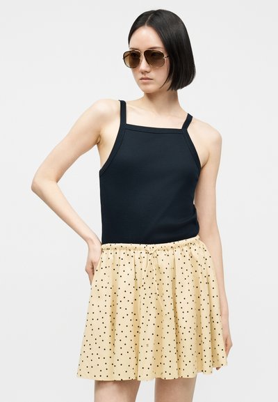 Γυναίκα με κοντά μαύρα μαλλιά που φοράει μεγάλα καφέ γυαλιά ηλίου, μαύρο αμάνικο τοπ και μπεζ φούστα με μαύρες πουά βούλες.