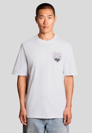 Jonge man draagt een wit T-shirt met een hartvormig "Lyle’s Laundry"-logo op de linkerborst, gecombineerd met lichtblauwe jeans.