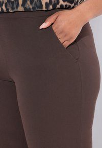 Bruna leggings gjorda av ett mjukt, elastiskt tyg, med en sidoficka och en åtsittande design, hålls i en hand.