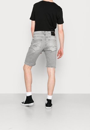 Homme portant un t-shirt noir, un short en jean gris clair, des chaussettes blanches et des baskets montantes noires, debout de dos sur un fond uni.