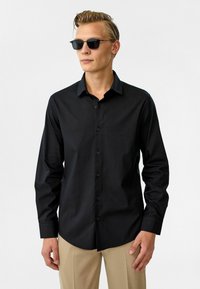 Zwarte lange mouw shirt met knopen, spitse kraag en een gladde textuur. Gekenmerkt door zwarte zonnebrillen en beige broeken.