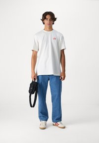 Camiseta de algodón blanca con un pequeño logo rojo, combinada con jeans de mezclilla azul claro y zapatillas blancas con rayas rojas. Lleva una bolsa negra.
