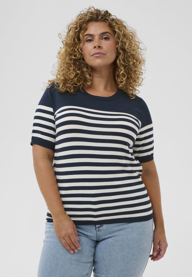 Donna con capelli biondi ricci che indossa una maglietta a maniche corte a righe orizzontali blu navy e bianco e jeans azzurri, in piedi davanti a uno sfondo semplice.