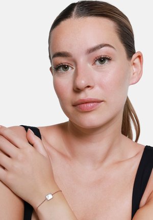 Silbernes Armband mit einem quadratischen, reflektierenden Mittelpunkt an einer zarten Kette, getragen an einem nackten Handgelenk, vor einem neutralen Hintergrund.