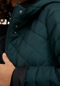 Chaqueta acolchada en azul petróleo oscuro con cuello alto, cierre de cremallera y botones de presión. Textura interior negra visible en el cuello y las mangas.