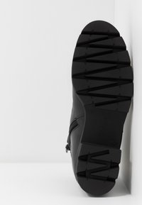 Semelle de botte noire avec des motifs de bande de roulement en zigzag profond et en blocs, reposant verticalement contre un mur d'angle blanc.