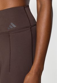 Leggings marron à taille haute avec une texture lisse, présentant des coutures contrastantes et un logo gris en haut à gauche.