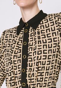 Gros plan d’un chemisier beige à motifs avec des formes géométriques noires, un col noir et des boutons noirs bordés d’or à l’avant.