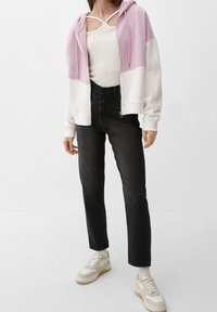 Felpa con zip di colore viola chiaro e bianco, indossata sopra un top aderente color crema, abbinata a jeans neri a vita alta e sneaker beige.