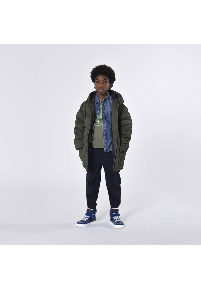 Giacca imbottita verde su una camicia di denim, abbinata a una t-shirt grafica verde, pantaloni scuri e sneakers alte blu.