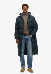Lange pufferjas in donker groen, met een gestructureerde afwerking en een capuchon. Gedragen over een beige turtleneck, gecombineerd met lichtblauwe jeans en beige schoenen.