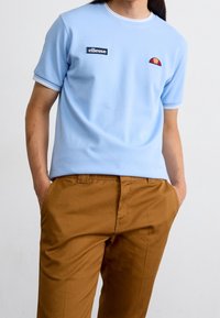 Jasnoniebieska koszulka z krótkim rękawem ozdobiona granatowymi i czerwonymi naszywkami z logo, z białym wykończeniem na kołnierzu i rękawach, zestawiona z brązowymi spodniami.