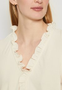 Cream top with a V-neck and ruffled edges. Soft fabric, featuring delicate ruffles along the neckline. Gold-tone earrings visible.