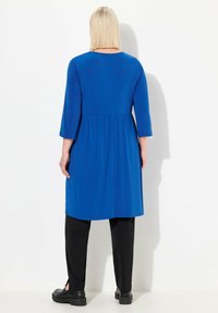 Niebieska sukienka do kolan z rękawami o długości trzech czwartych, luźny krój i szefowym paskiem w talii, w połączeniu z czarnymi spodniami i masywnymi czarnymi butami.