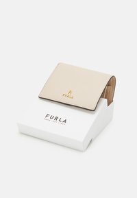 Peněženka Furla v světle béžové kůži s texturovaným povrchem, s zlatým logem a skládacím designem, umístěná na bílé krabičce.