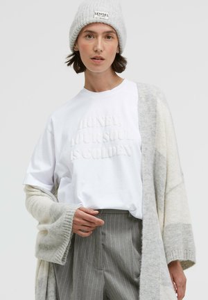 White t-shirt with embossed text, grey and white oversized cardigan, and grey pinstripe trousers. Soft textures, relaxed fit. Light beanie.