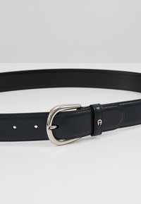 Cinturón de cuero azul marino con hebilla de metal plateado, textura suave, ajustable con varios agujeros y un pequeño acento de marca.