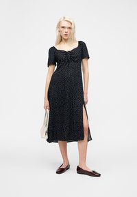 Femme blonde portant une robe midi noire à pois blancs, avec une fente latérale, tenant un sac à main blanc, et portant des chaussures plates marron foncé.