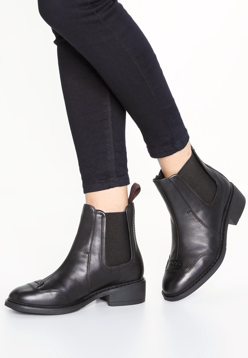 Personne portant des bottines Chelsea en cuir noir et un pantalon slim foncé, debout sur une surface blanche avec un pied légèrement soulevé.