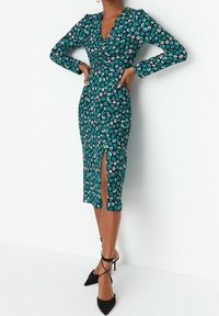 Femme portant une robe midi florale teal avec des manches longues, une fente à l'avant et des talons noirs à bout pointu avec des brides autour des chevilles.