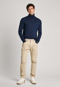 Jūras spēku turtlenecks džemperis, bēša krāsas chinos bikses, baltas sneakers ar gaiši brūniem zole. Džemperim ir cieša piegriezne un pieguļoši piedurknes. Vienkāršs dizains.