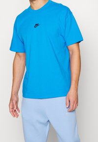 Blå bomullströja med korta ärmar och rundad halsringning, med en svart Nike-logga på bröstet, kombinerad med ljusblå byxor.