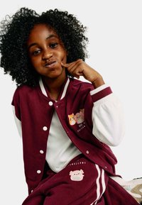 Burgundská bunda s bílými rukávy, zdobená výšivkou zvířat; spárována s odpovídajícími burgundskými kalhotami se bočními pruhy a grafikou.