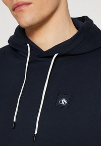 Sudadera azul marino con cordones blancos, textura acanalada y un pequeño parche con logo bordado en el pecho.