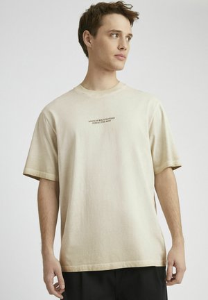 T-shirt en coton beige avec une coupe décontractée, des manches courtes et un col rond. Contient un texte imprimé en marron foncé sur la poitrine.