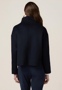 Abrigo corto de color navy con un cuello alto, hecho de una tela suave. Presenta hombros caídos y un diseño de líneas limpias, combinado con pantalones oscuros.
