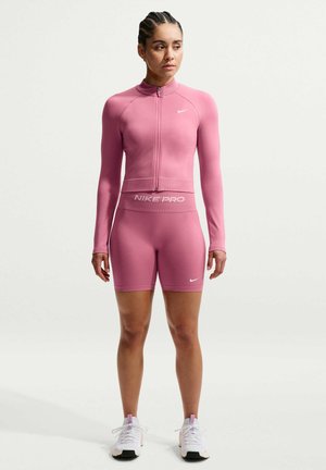 Frau mit geflochtenem Haar, die ein pinkes Nike Pro Zip-up Langarmshirt, passende Shorts und weiße Sneakers trägt und vor einem schlichten Hintergrund steht.