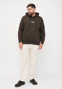 Hoodie en coton marron avec une poche avant et un capuchon à cordon, associé à un pantalon beige clair et des chaussures noires à lacets.